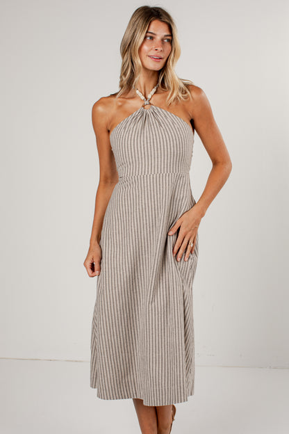 A woman modeling a halter-neck midi dress with vertical beige and white stripes, featuring a knotted neckline and side pockets, set against a plain light gray studio background.