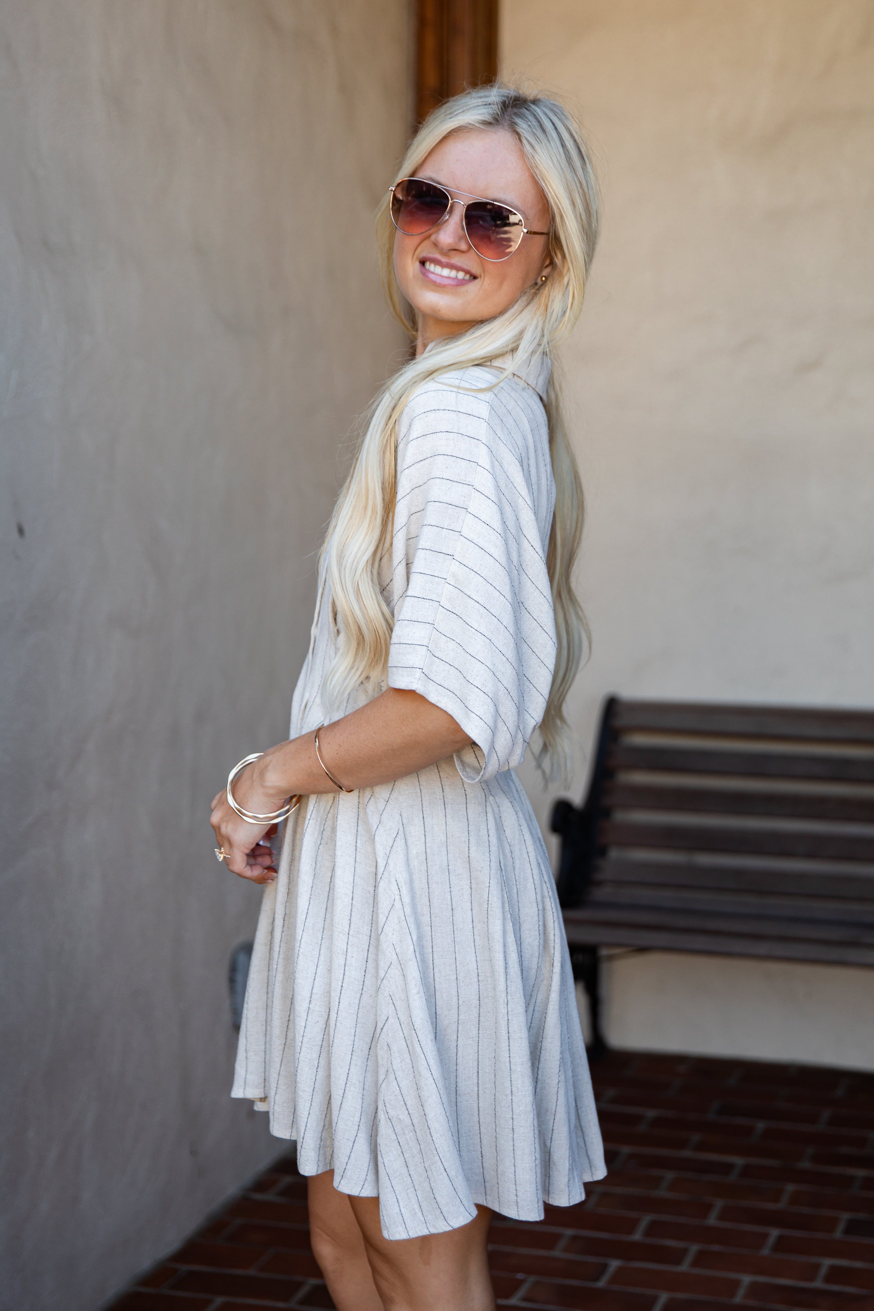 A woman with long blonde hair and sunglasses wearing a white short-sleeve dress with thin vertical blue stripes, standing in an outdoor setting with a stucco wall and brick floor.