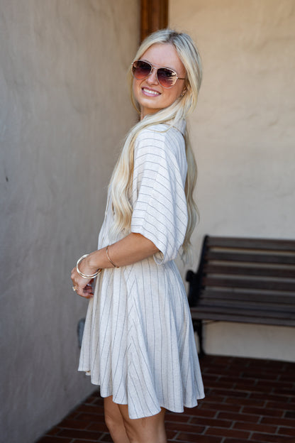 A woman with long blonde hair and sunglasses wearing a white short-sleeve dress with thin vertical blue stripes, standing in an outdoor setting with a stucco wall and brick floor.