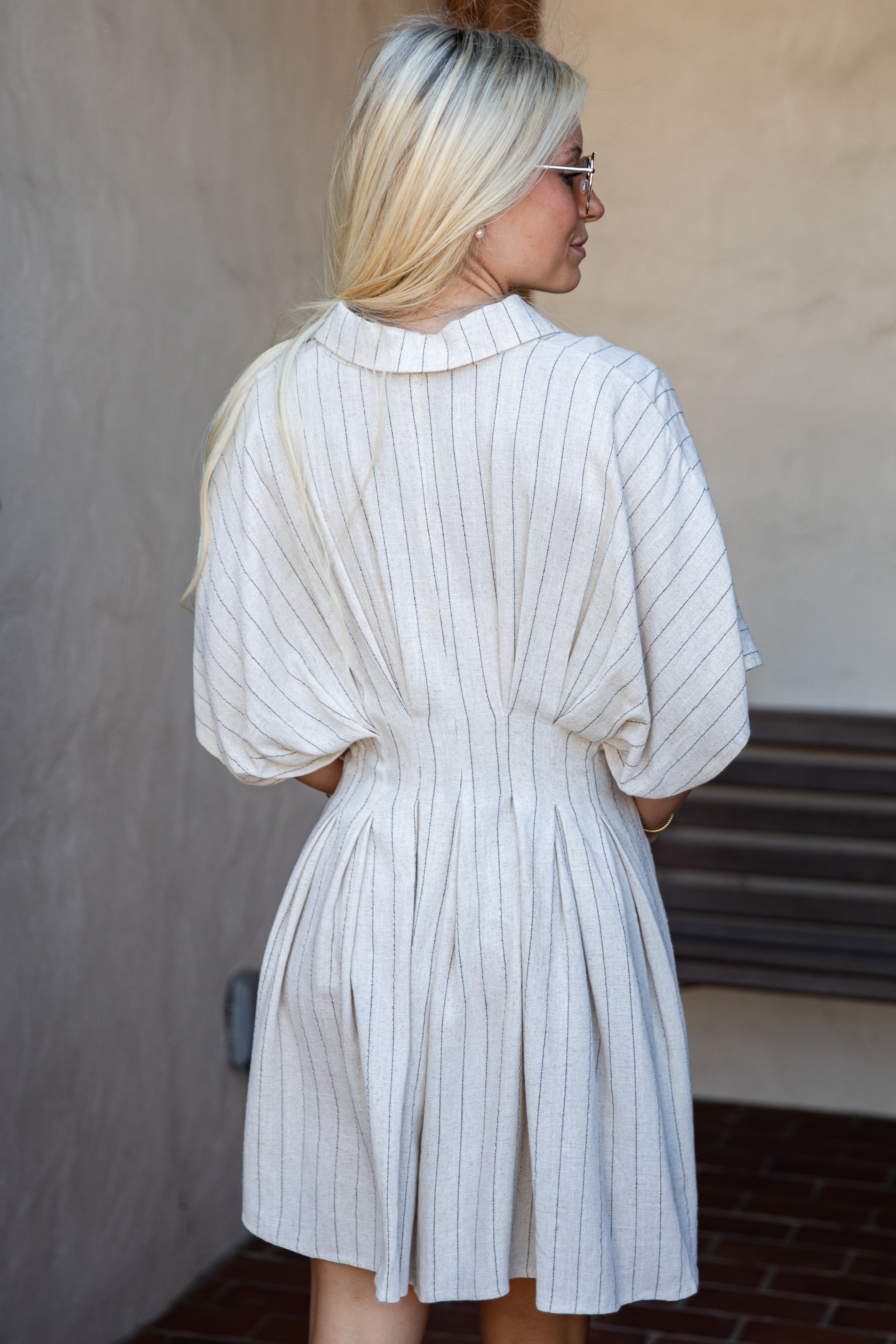 A woman with blonde hair is shown from behind wearing a cream-colored pleated shirt dress with thin vertical blue stripes. She is standing outdoors against a textured beige wall.