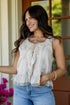 A woman wearing a sleeveless cream-colored blouse with ruffles and lace trim, paired with light blue jeans, standing in front of a porch window.