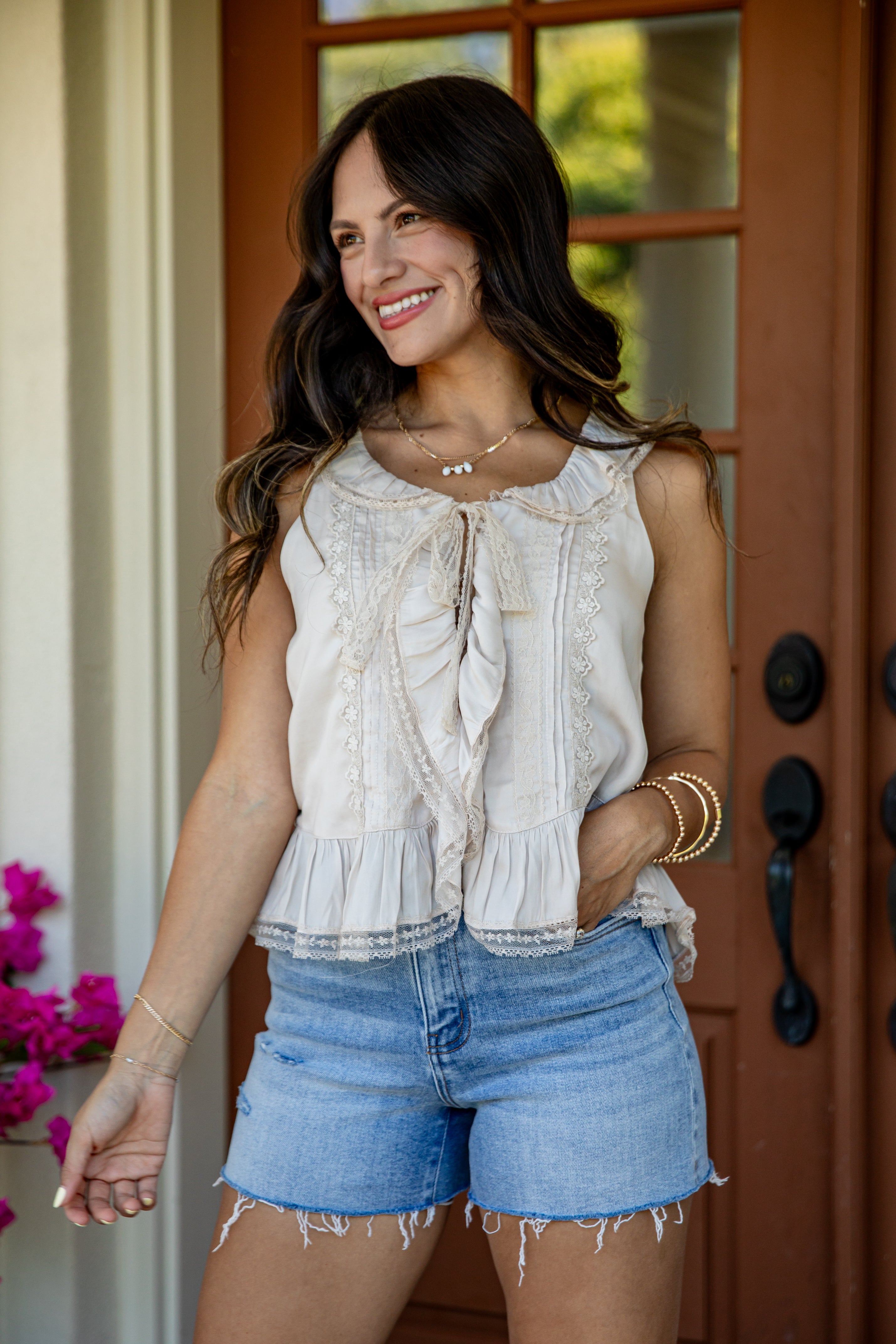 A woman wearing a sleeveless cream ruffled blouse and light blue denim shorts, standing in front of a brown wooden door.