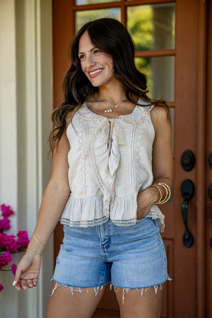 A woman wearing a sleeveless cream ruffled blouse and light blue denim shorts, standing in front of a brown wooden door.