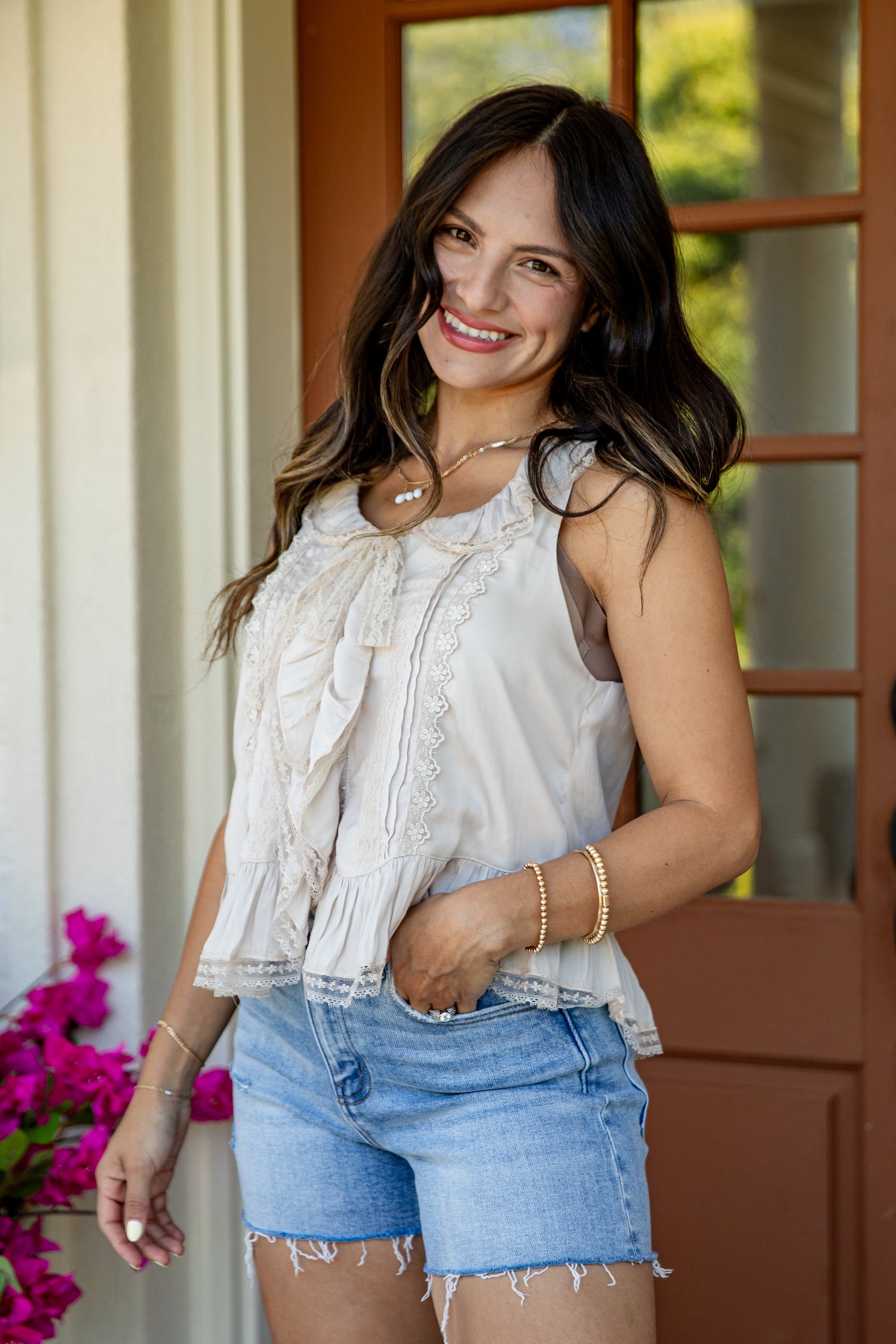 A woman wearing a sleeveless cream ruffled blouse and light blue denim shorts, standing in front of a brown wooden door with greenery visible in the background.