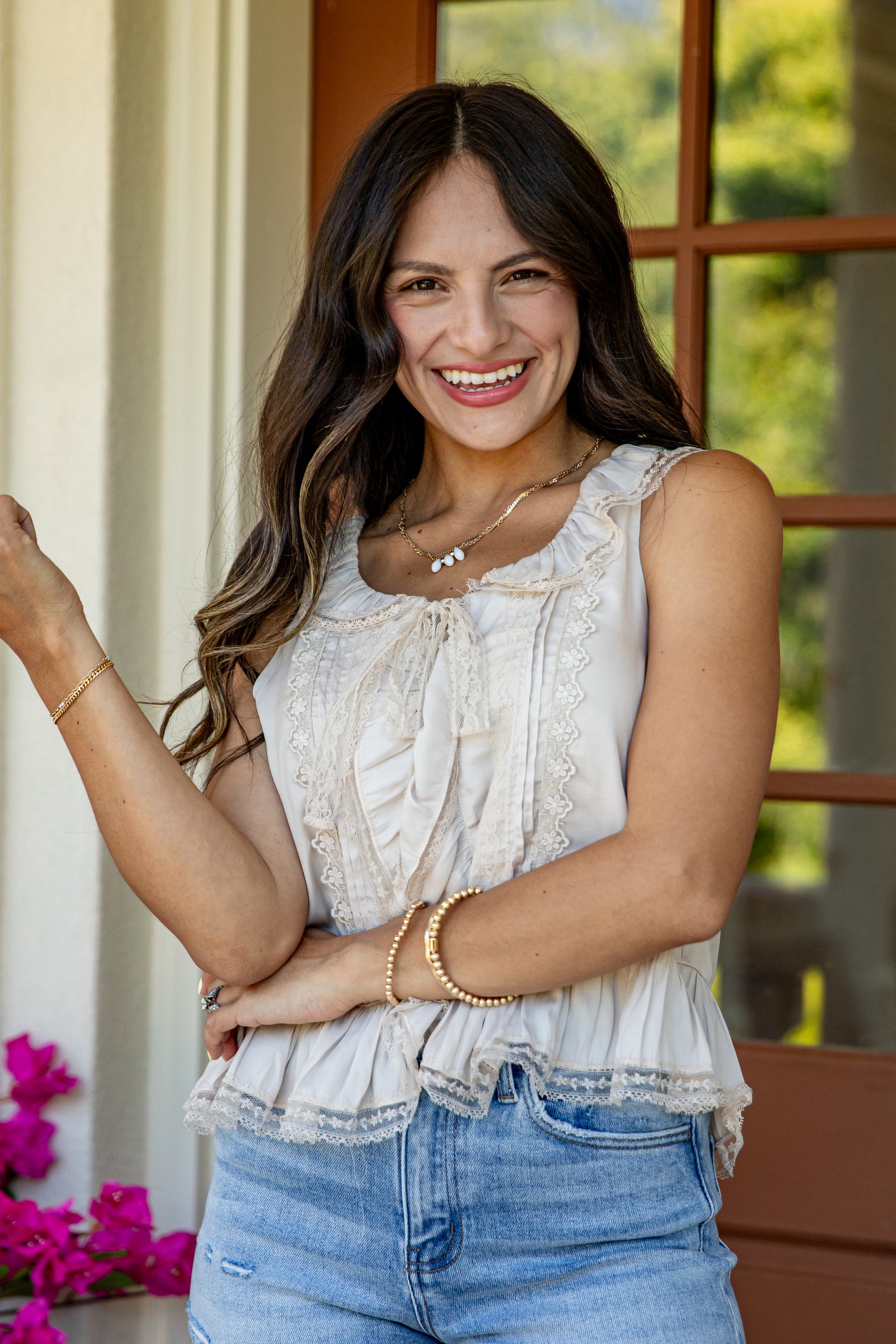 A smiling woman with long dark brown hair wearing a sleeveless cream ruffled blouse and light blue jeans, standing in front of a window with warm wood trim.