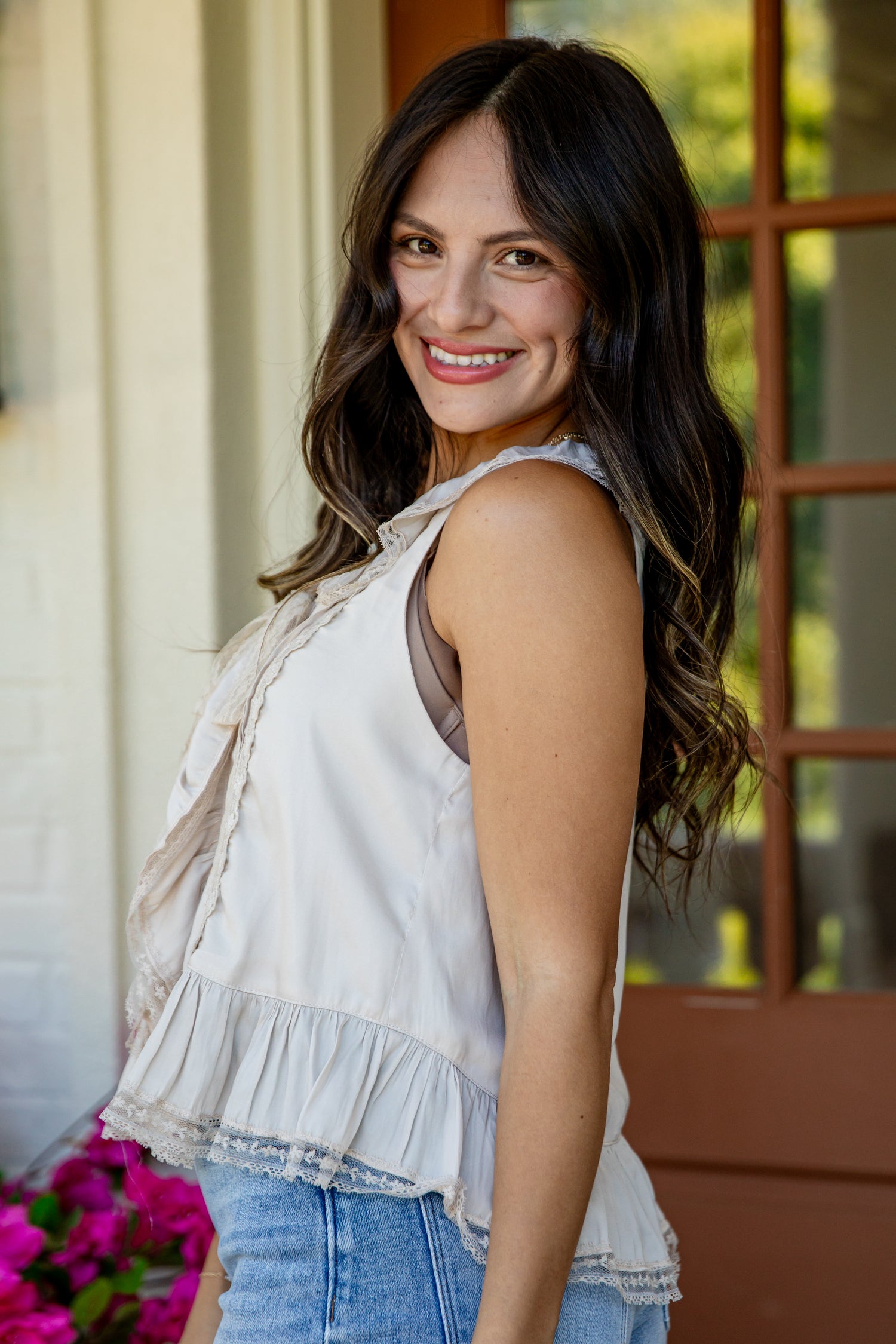 A woman with long dark hair smiling and looking back over her shoulder, wearing a sleeveless ruffled cream top and light blue jeans, standing in front of a porch door.