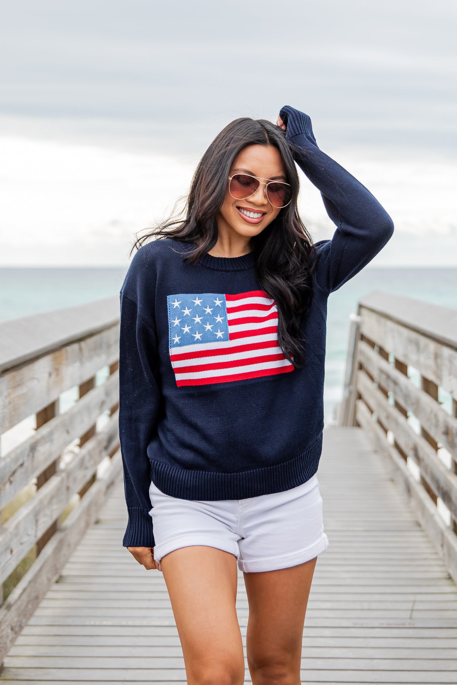 Woman wearing a navy sweater with an American flag design on a wooden pier by the ocean.