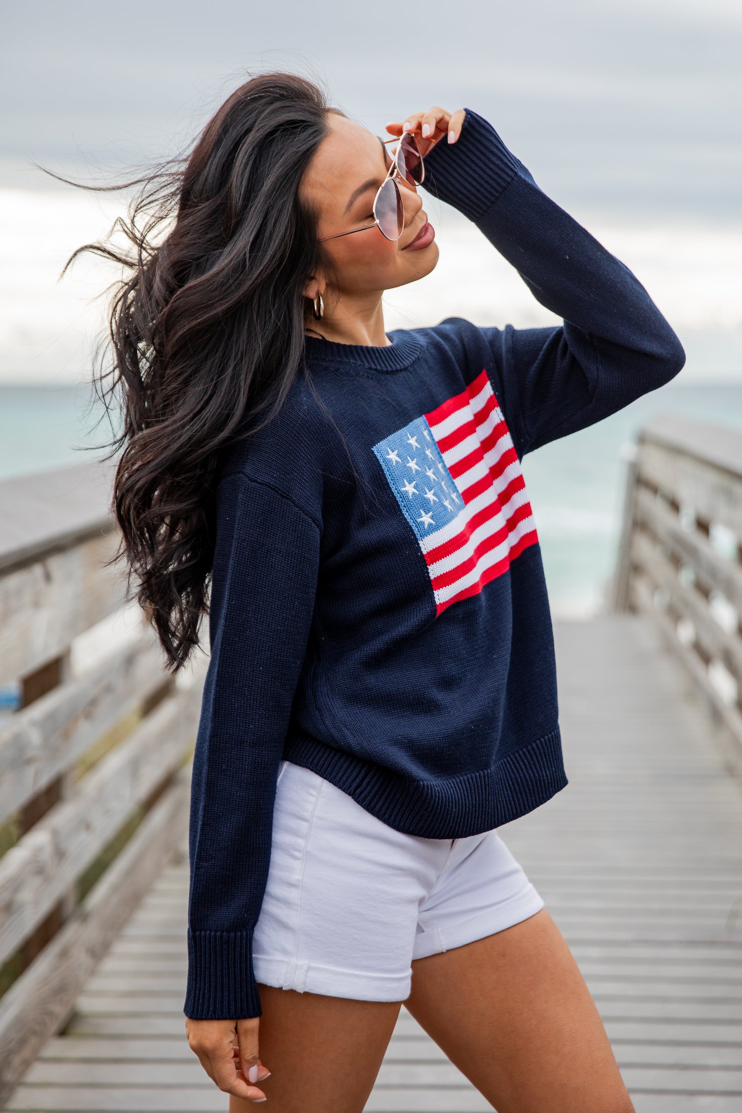 Woman wearing a navy sweater with an American flag design on a wooden pier by the ocean.
