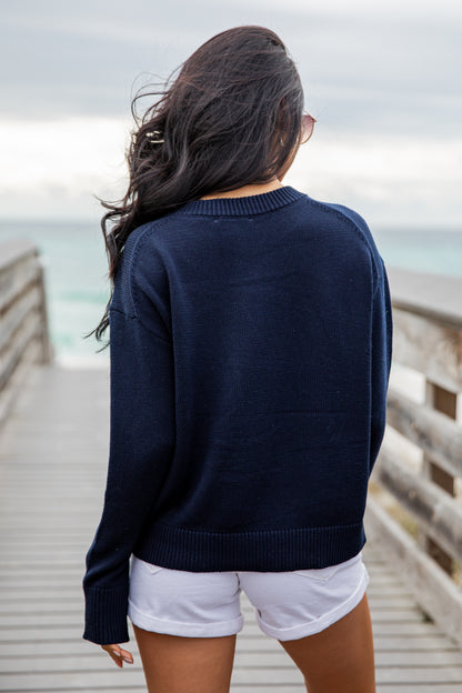 Woman wearing a navy sweater with an American flag design on a wooden pier by the ocean.