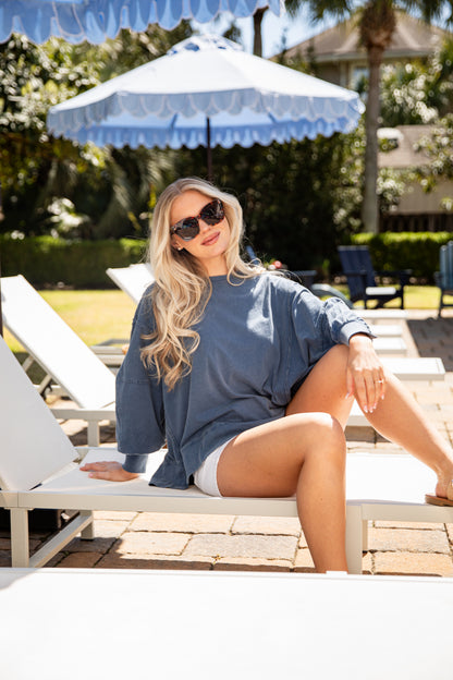 Woman sitting on a lounge chair under a blue and white umbrella in a garden setting.