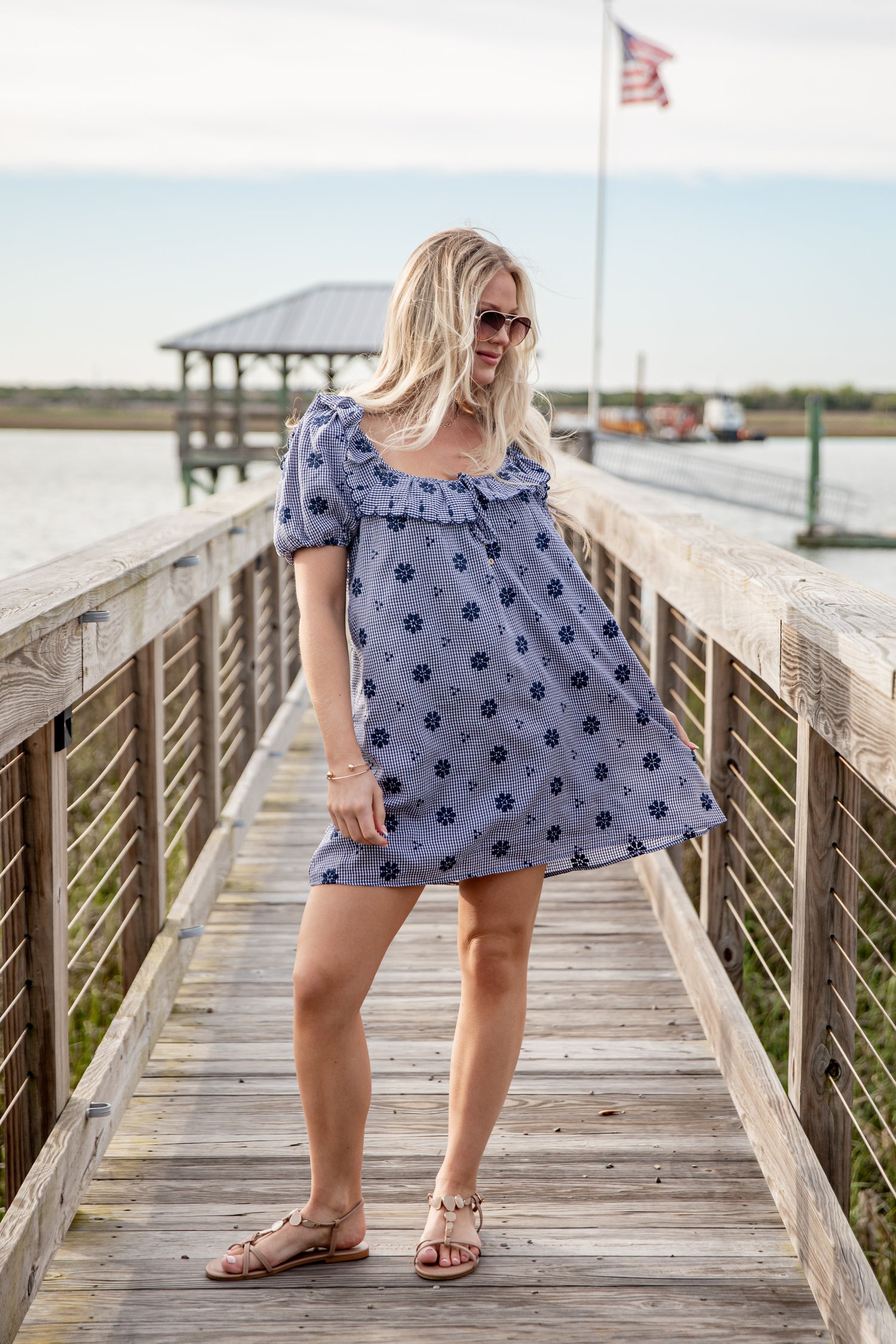 Woman in a blue floral dress standing on a wooden dock by a body of water.