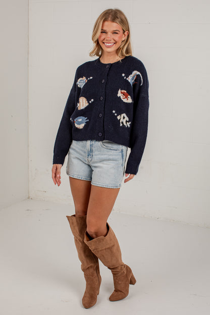 A woman modeling a navy blue knit cardigan with white, red, and blue fish graphics, paired with light-wash denim shorts and brown suede boots, standing against a white studio wall.