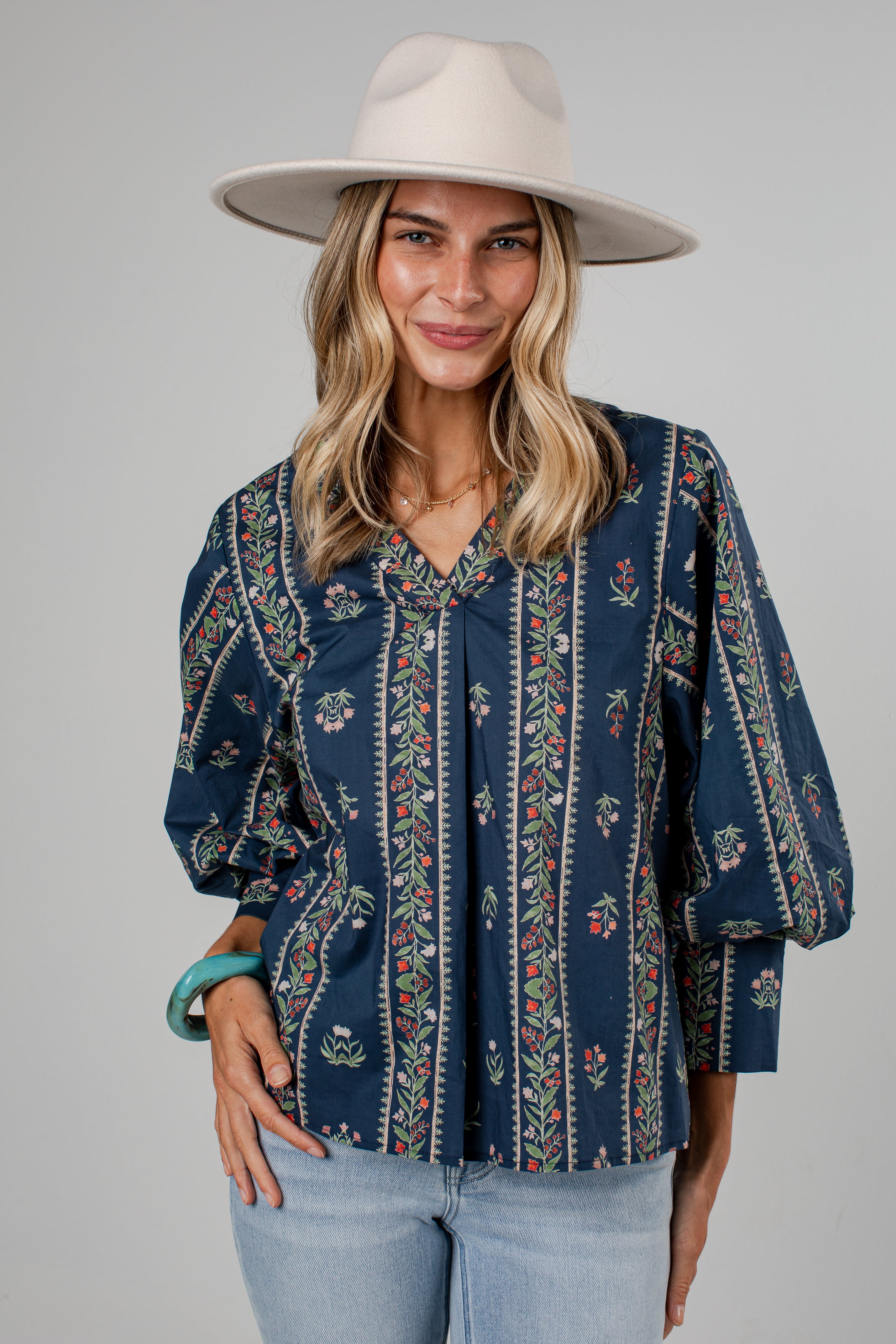 A woman wearing a navy blue bohemian-style blouse with vertical floral patterns, light blue jeans, and a wide-brimmed cream hat, photographed against a solid gray studio background.