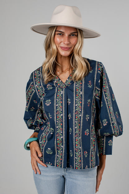A woman wearing a navy blue bohemian-style blouse with vertical floral patterns, light blue jeans, and a wide-brimmed cream hat, photographed against a solid gray studio background.