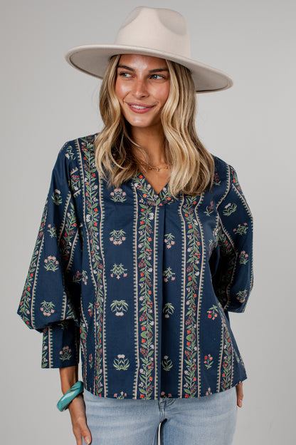 A woman wearing a navy blue bohemian-style blouse with vertical floral and leaf patterns, paired with light blue jeans and a beige wide-brim hat, against a solid gray studio background.