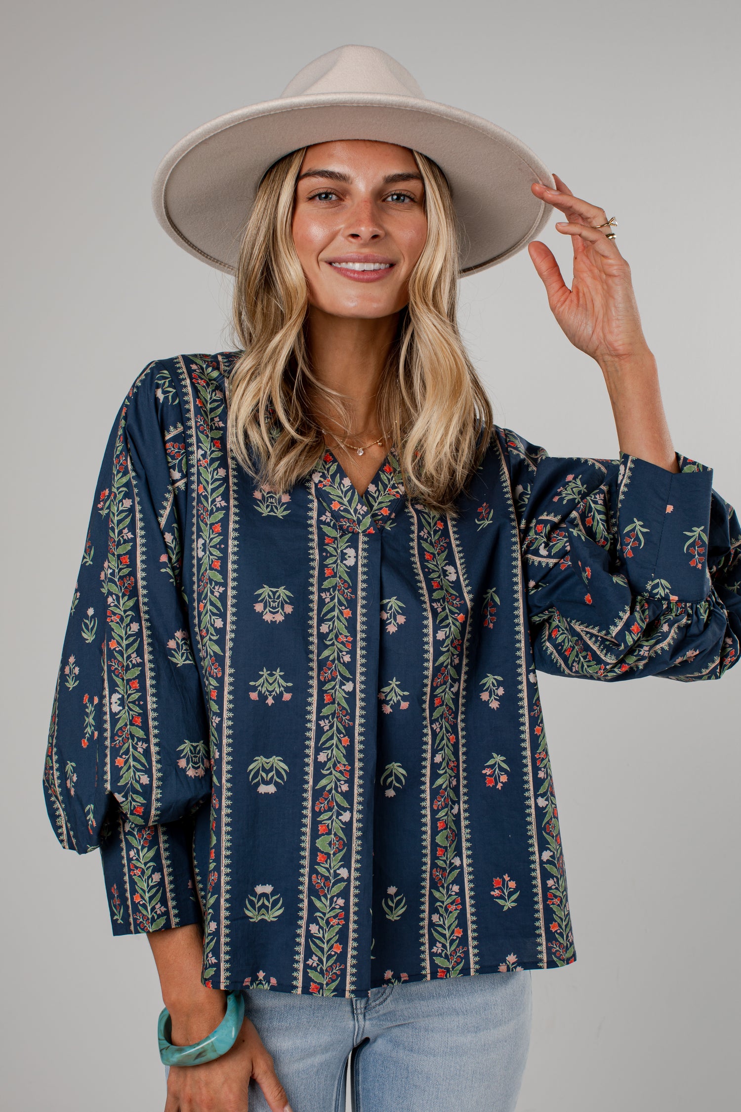 A woman wearing a navy blue bohemian-style blouse with vertical floral patterns, paired with a wide-brimmed beige hat and light blue jeans, photographed against a solid gray studio background.