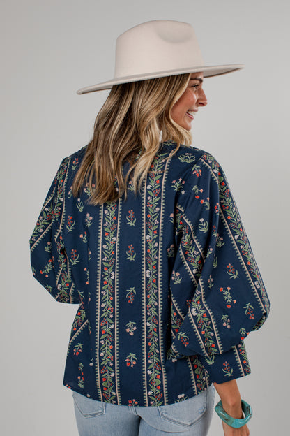 A woman wearing a navy blue bohemian-style blouse with vertical floral and geometric patterns, paired with a wide-brimmed cream hat and light blue jeans, shown from a three-quarters back view against a solid gray background.