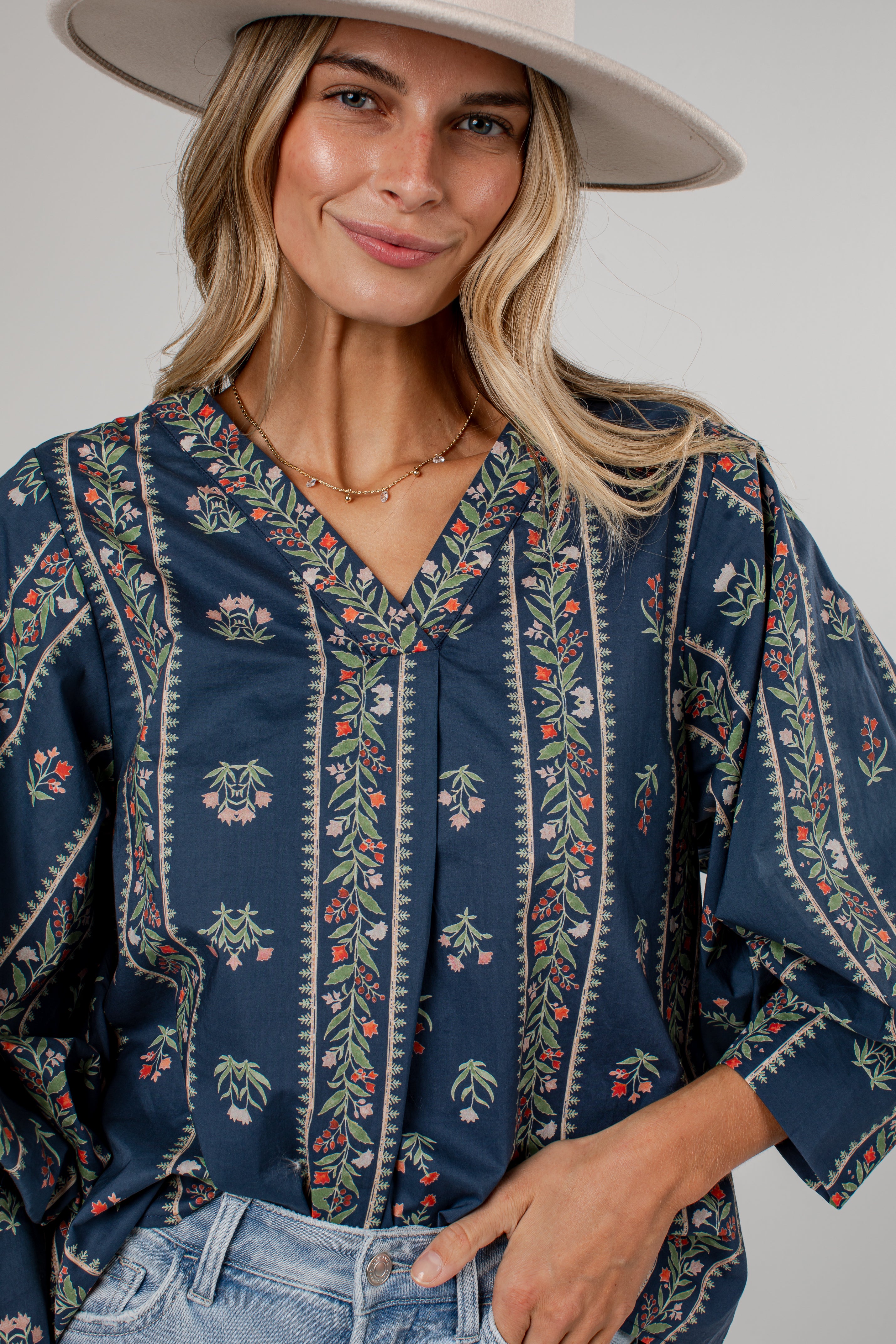 A woman wearing a navy blue bohemian-style blouse with colorful floral embroidery and vertical patterned borders, paired with light-wash jeans and a wide-brimmed beige hat against a neutral gray background.