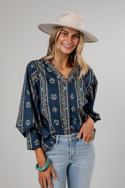 A woman wearing a navy blue bohemian-style blouse with vertical floral and geometric patterns, paired with a wide-brimmed cream hat and light blue jeans, shown from a three-quarters back view against a solid gray background.