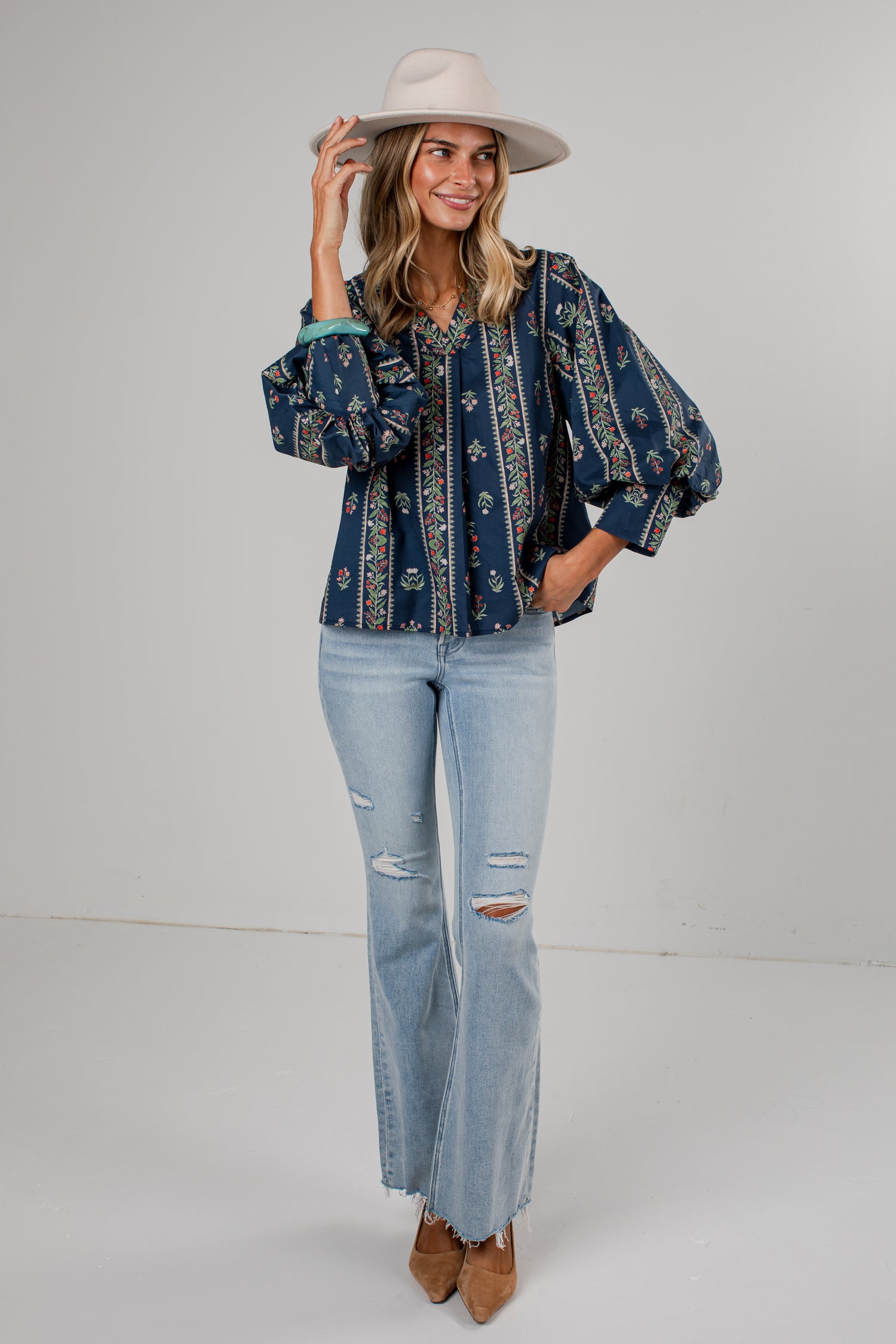 A woman modeling a bohemian-style navy blue blouse with intricate multicolor embroidery, paired with light-wash distressed jeans, a beige wide-brim hat, and brown boots, set against a plain light gray studio background.