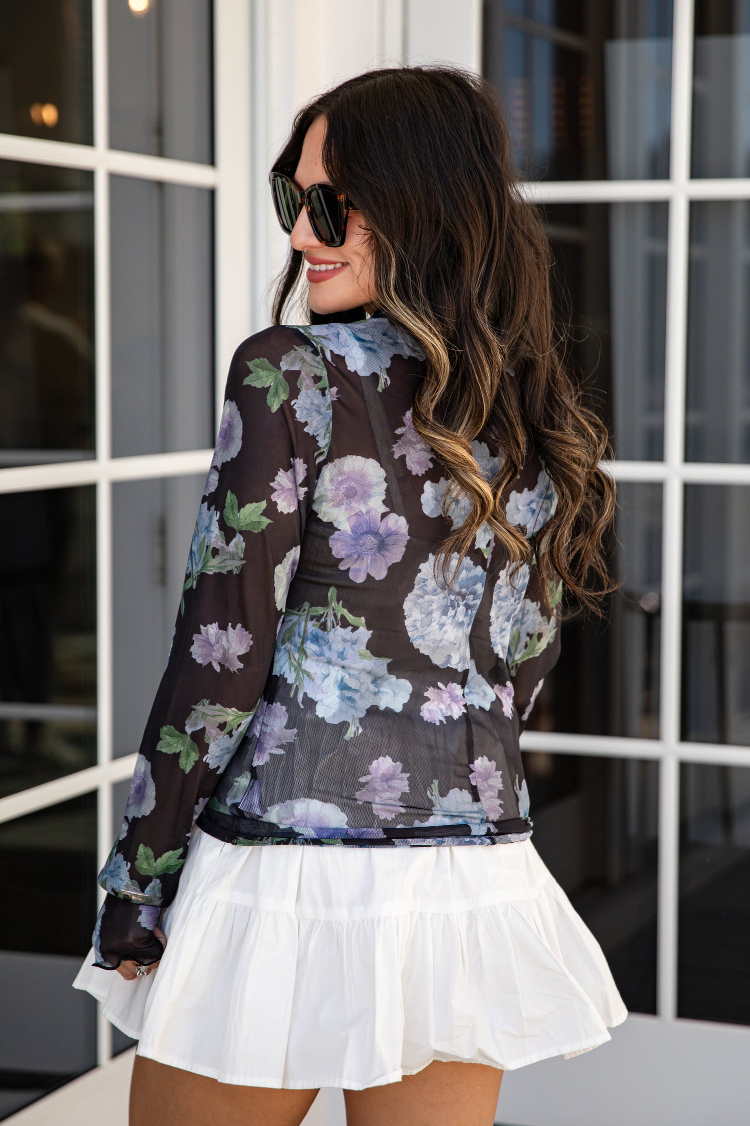 Woman wearing a floral blouse and white skirt in front of a window.