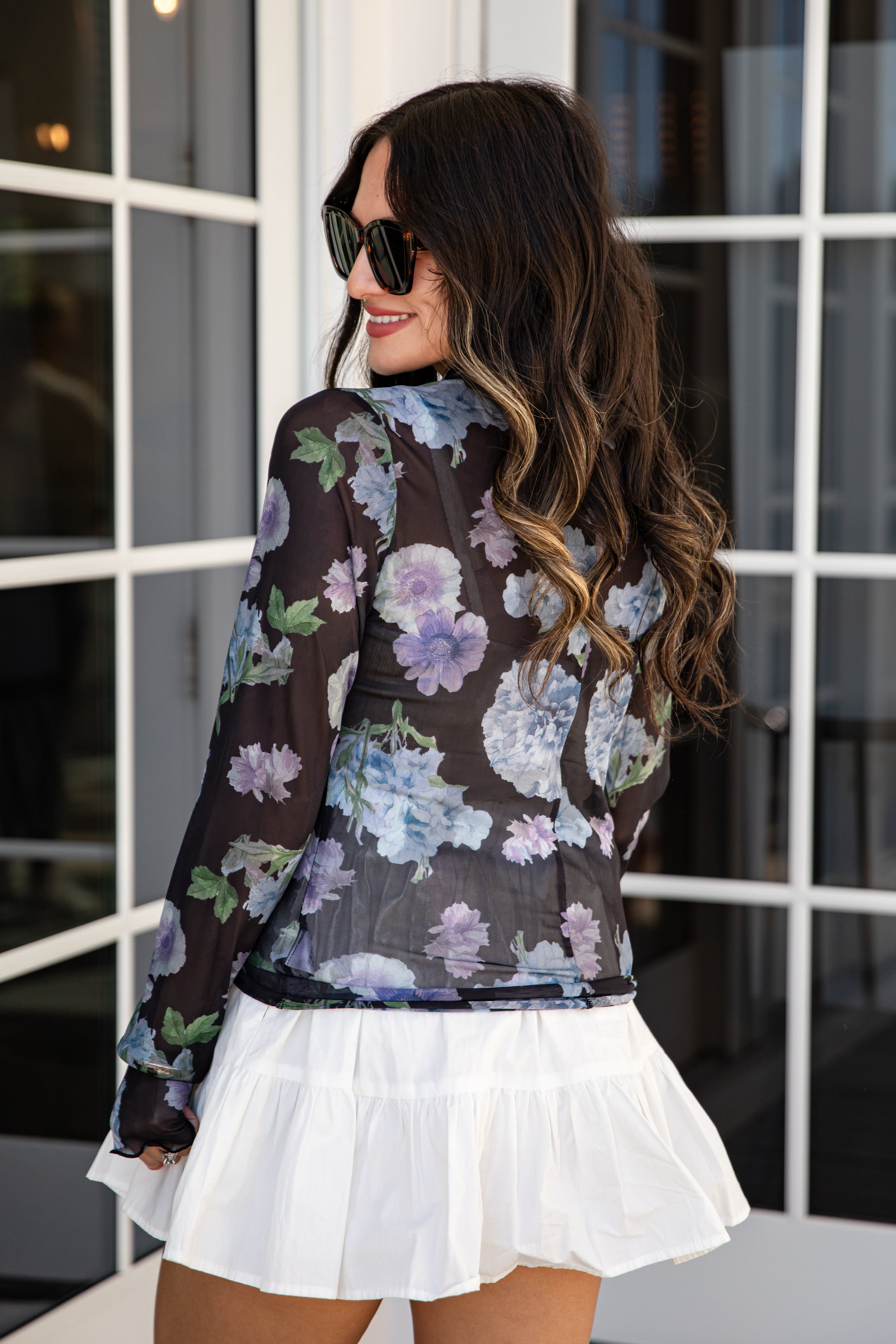 Woman wearing a floral blouse and white skirt in front of a window.