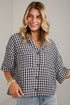 A woman wearing a navy and white gingham button-up shirt with three-quarter sleeves, paired with light blue jeans, standing against a light gray paneled wall.