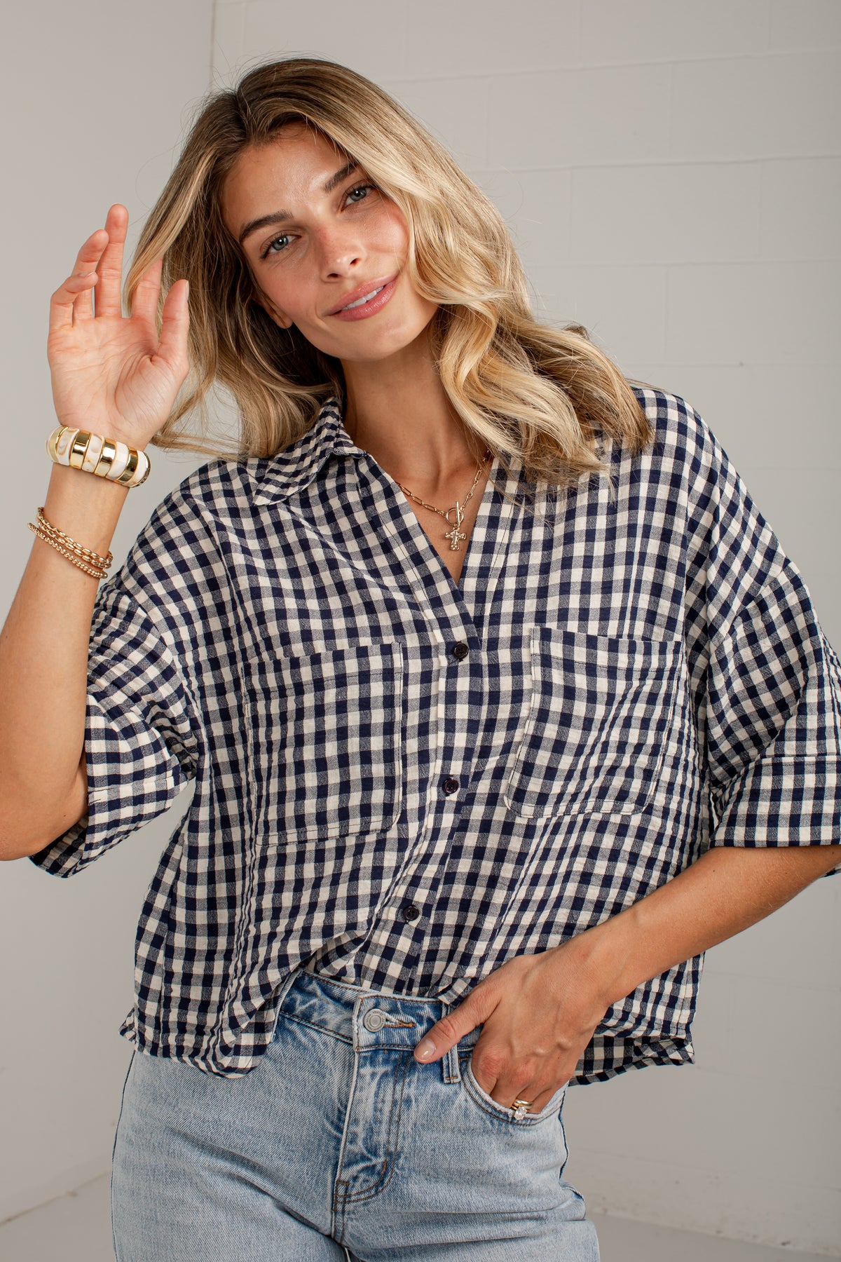 A woman wearing a navy and white gingham short-sleeve button-up shirt with light blue jeans, posing against a light gray paneled wall.