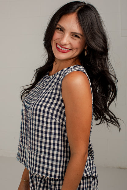 A smiling woman with long dark hair wearing a sleeveless navy and white gingham check top, photographed against a plain light gray background.