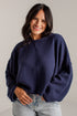 A woman wearing a navy blue textured knit sweater and light-wash jeans, standing against a light gray wall.