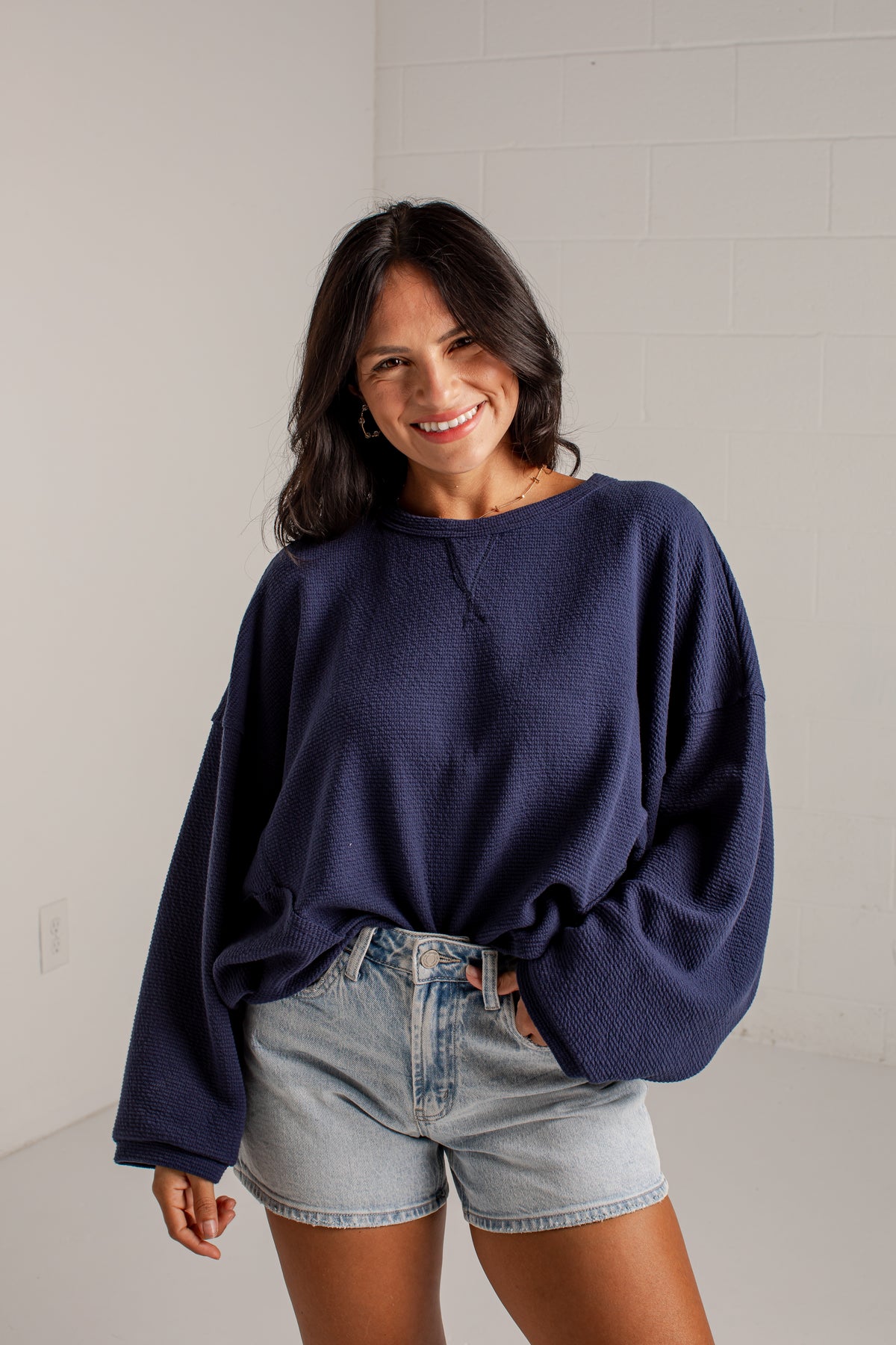 A woman wearing a dark navy blue knit crewneck sweater and light-wash denim shorts, standing against a white brick wall.