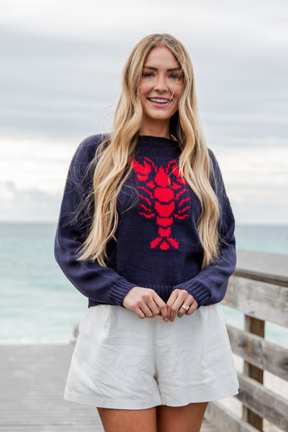 Woman wearing a navy sweater with a red lobster design by the ocean.