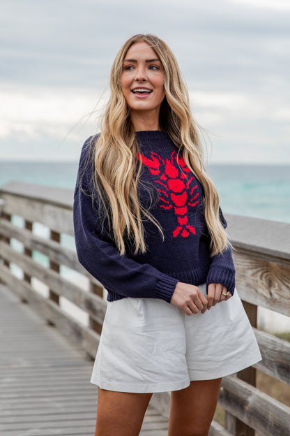 Woman wearing a navy sweater with a red lobster design on a wooden boardwalk by the ocean.