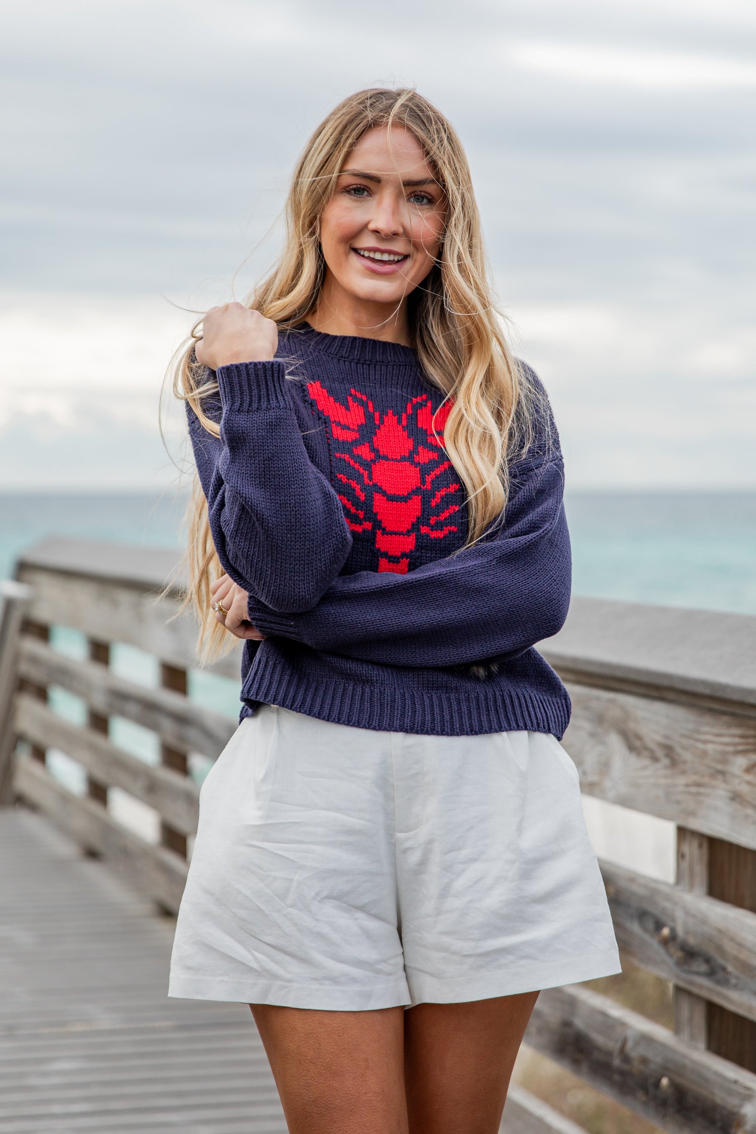 Woman wearing a navy sweater with a red lobster design and light shorts on a wooden pier.