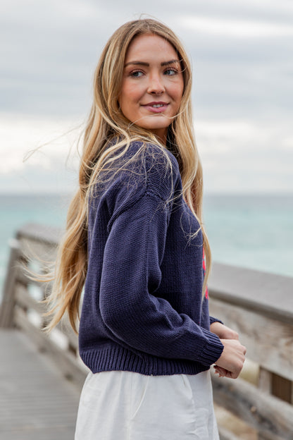 Woman wearing a navy sweater and white pants standing on a wooden pier with a blurred background