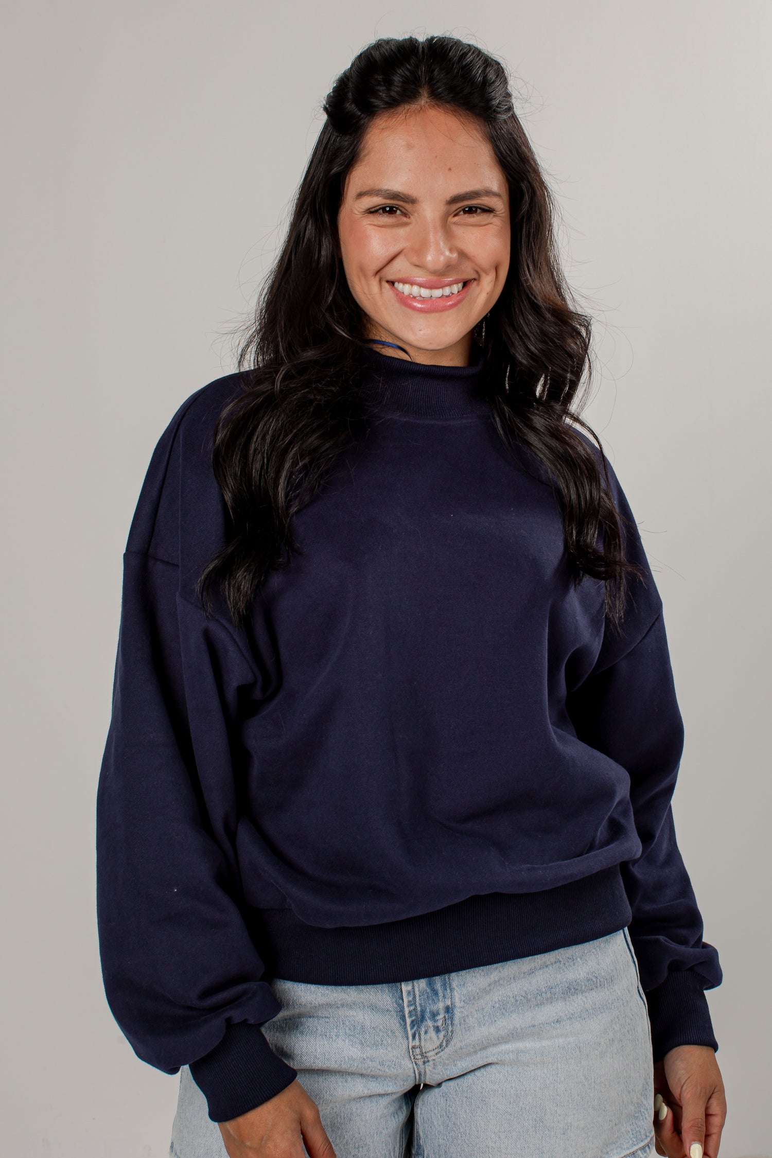 A smiling woman with long dark hair wearing a navy blue mock neck sweatshirt and light wash jeans, standing against a plain light gray studio background.