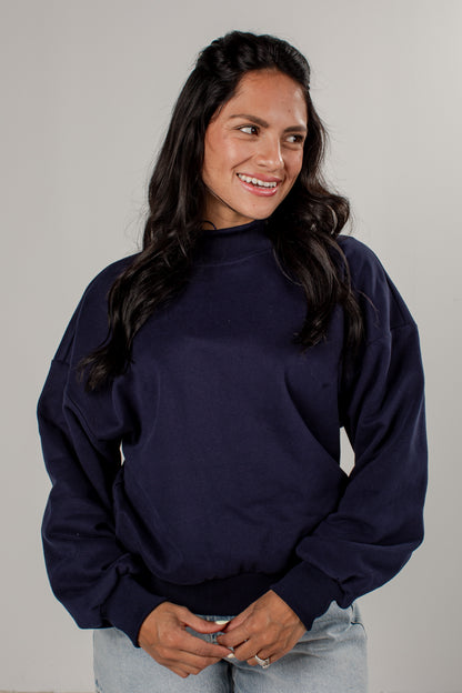 A woman with long dark hair wearing a navy blue mock neck sweatshirt and light wash jeans, standing against a plain light gray studio background.