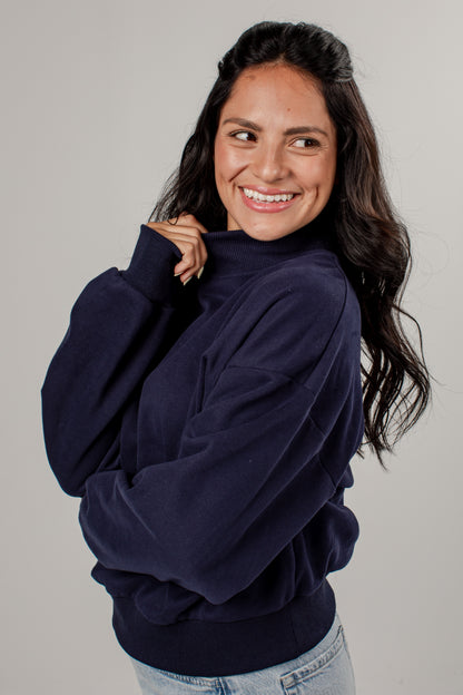 A woman with long dark hair smiling and looking to the side, wearing a dark navy blue mock-neck sweatshirt and light blue jeans, photographed against a solid light gray background.