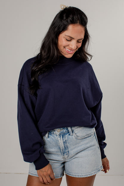 A woman wearing a dark navy blue crewneck sweatshirt and light-wash denim shorts, standing against a plain light gray studio background.