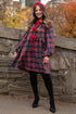Woman in a plaid dress with red accents standing against a stone wall with autumn trees and city buildings in the background.