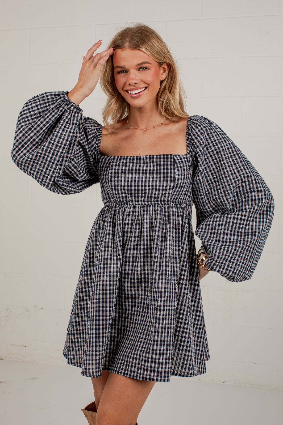 A woman modeling a navy and white gingham mini dress with voluminous puff sleeves, a square neckline, and a smocked waist, set against a white brick wall.