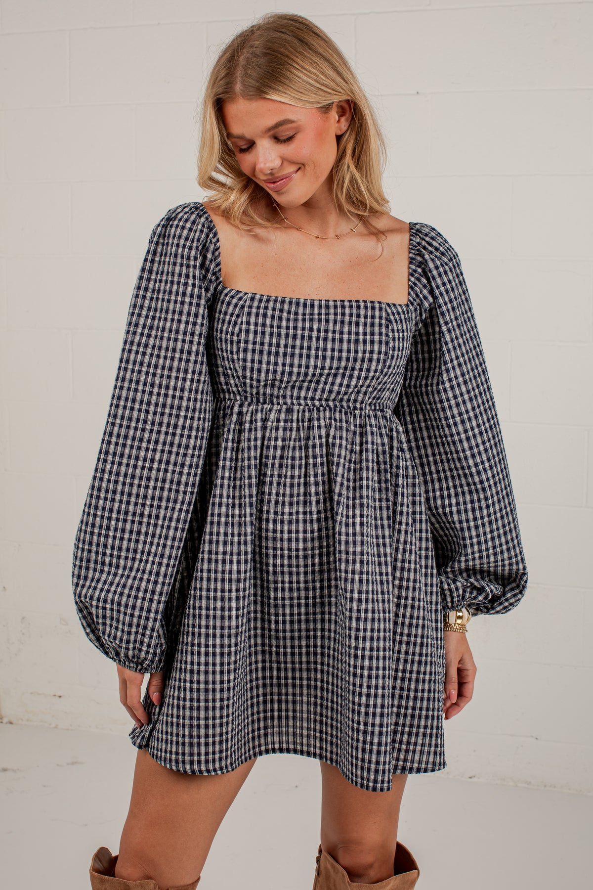 A woman wearing a navy and white gingham mini dress with puff sleeves and a square neckline, standing against a white brick wall.