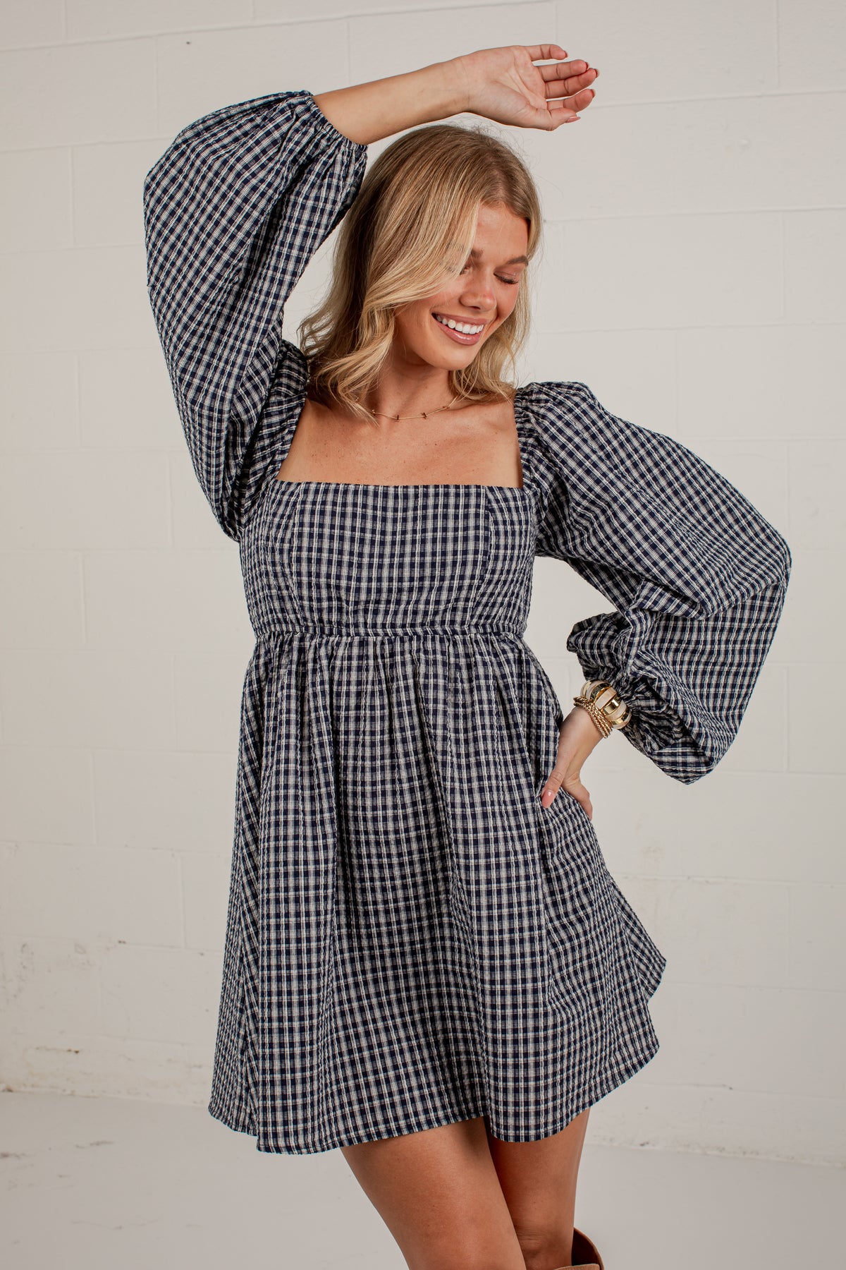 A woman modeling a navy and white gingham mini dress with voluminous puff sleeves and a square neckline, posing against a white paneled wall.