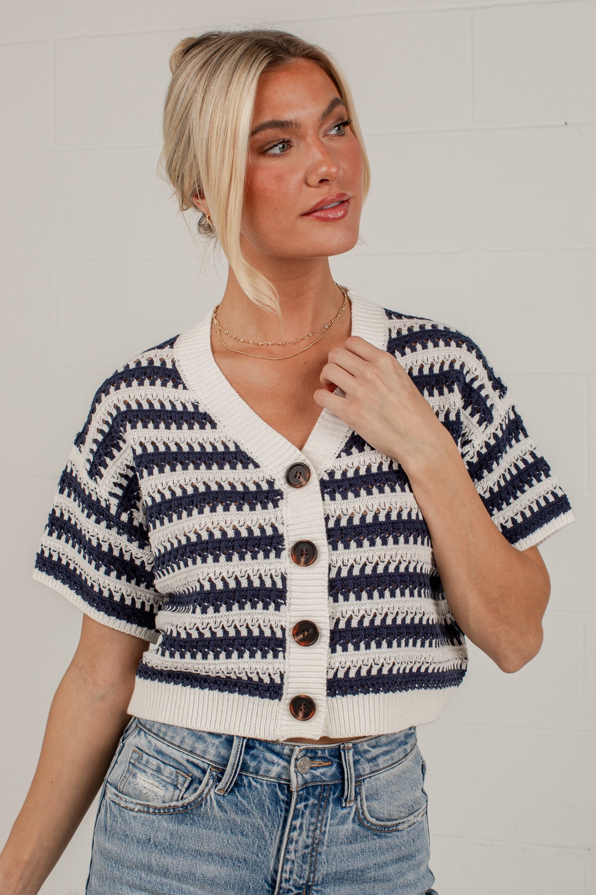 A woman wearing a short-sleeve knit cardigan with navy and cream horizontal stripes, brown buttons, and a V-neck, paired with blue jeans against a white brick wall.