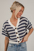 A woman wearing a short-sleeve knit cardigan with a navy and cream horizontal pattern, brown buttons, and blue jeans, standing against a white brick wall.