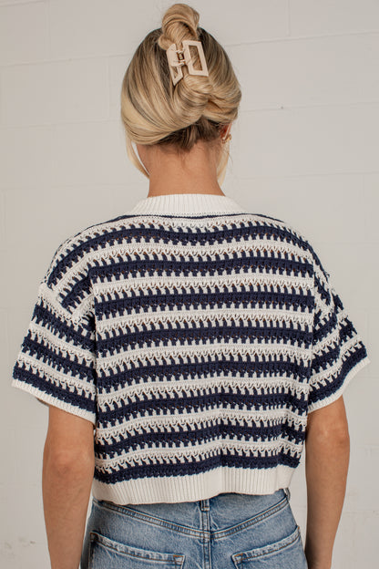 A woman viewed from behind wearing a short-sleeve knit top with horizontal navy and off-white stripes, paired with blue jeans against a white paneled wall.