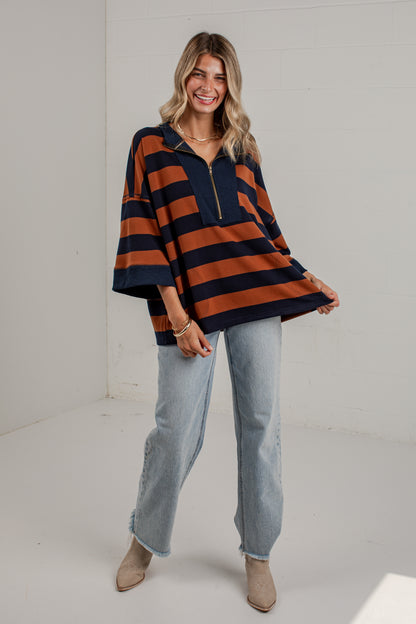 A woman modeling a loose, oversized short-sleeve top with bold horizontal navy and rust-orange stripes, paired with light-wash jeans and beige ankle boots, in a bright white studio setting.