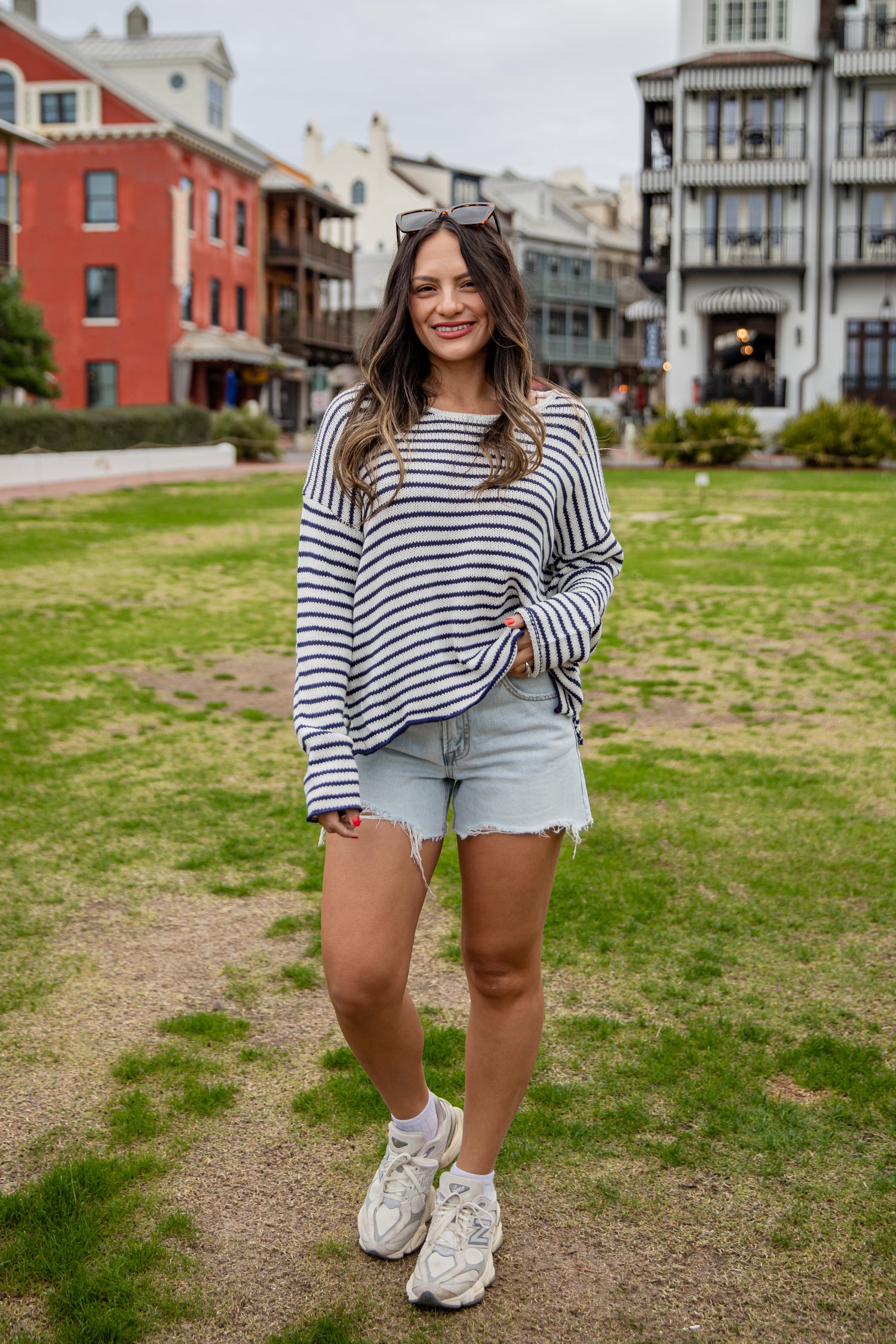 Woman in a striped sweater and shorts standing on a grassy area with buildings in the background