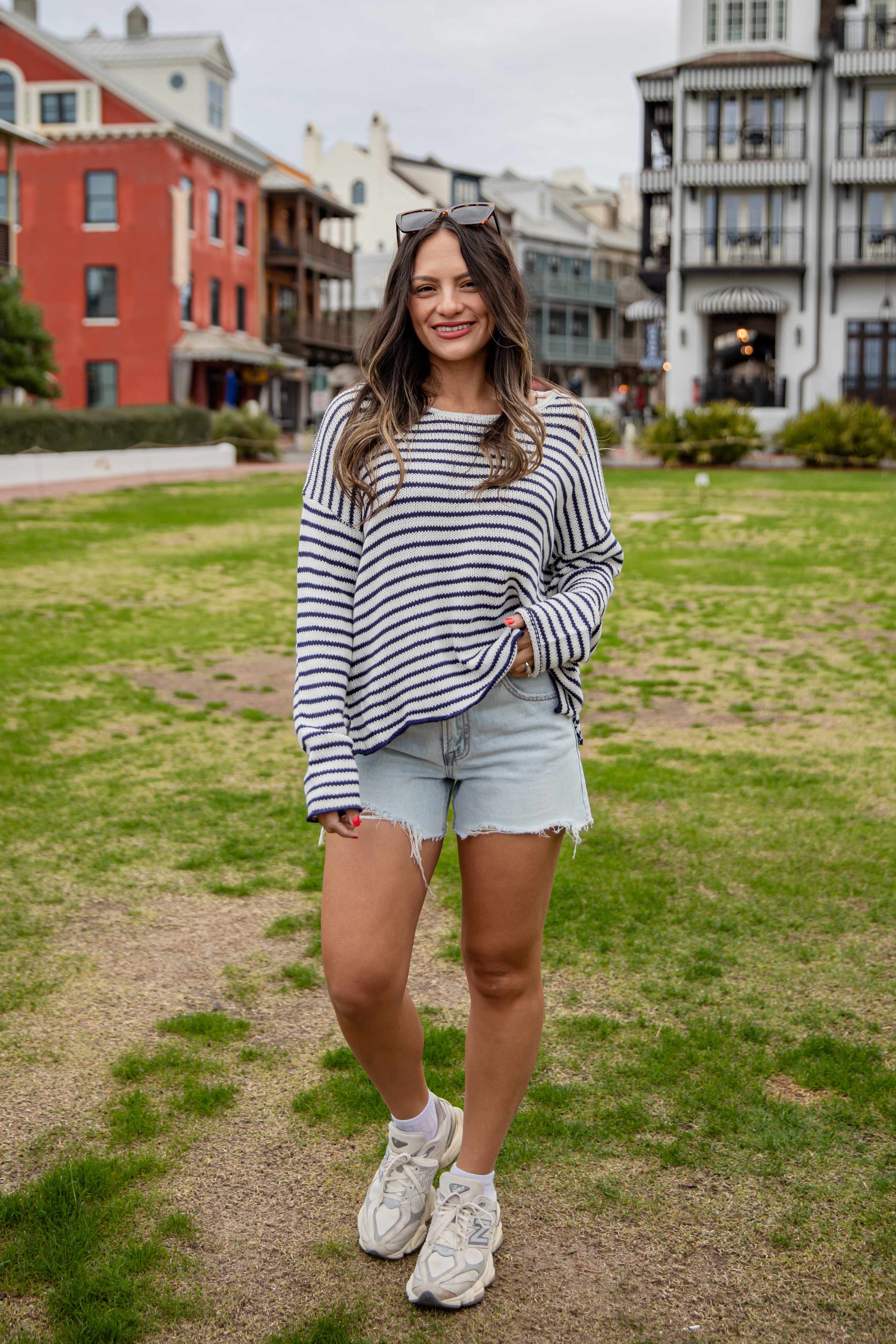 Woman in a striped sweater and shorts standing on a grassy area with buildings in the background