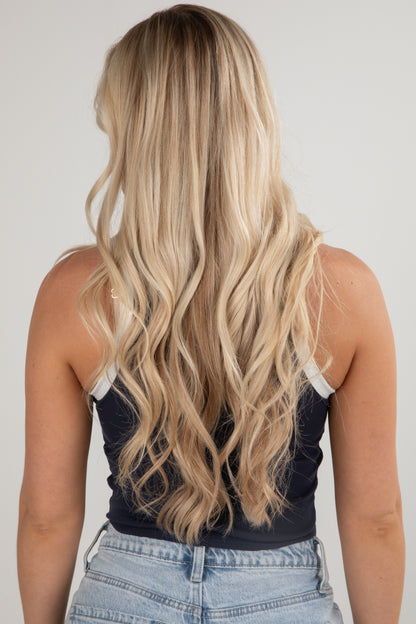 Person with long, wavy blonde hair wearing a dark tank top and light blue jeans against a plain background